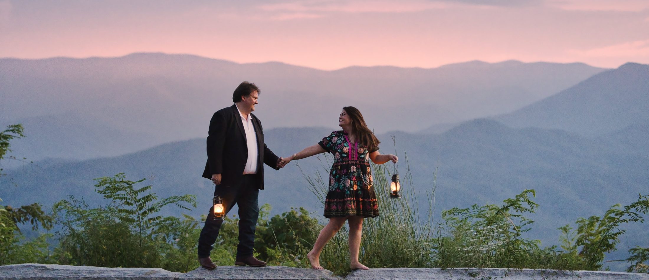 The two of us holding hands and lanterns on a stone wall at a mountain overlook, pink sunset over the ridges behind us